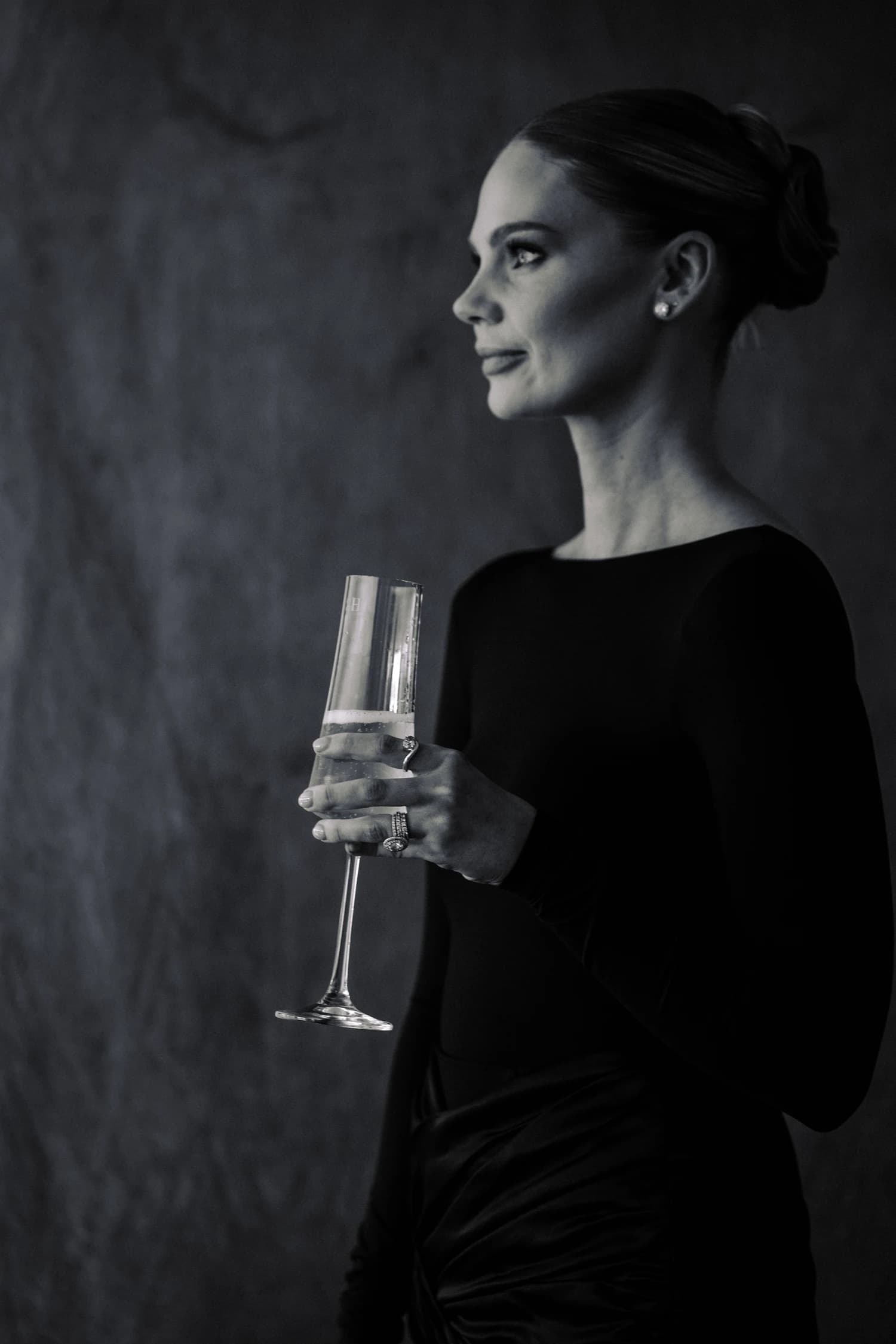 A woman in a long-sleeved black gown holding a champagne flute against a dark textured wall.