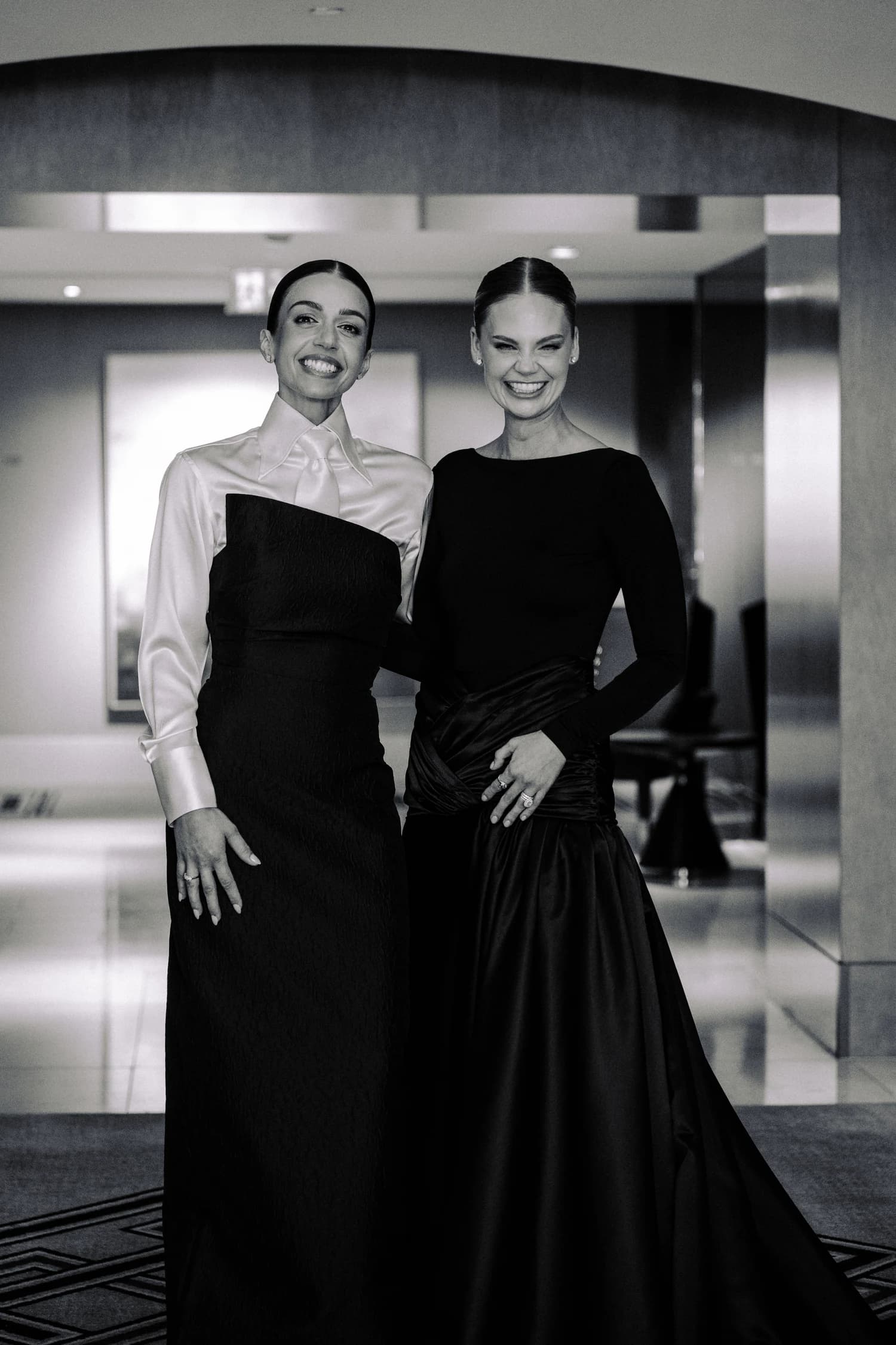 Two women in eveningwear standing together, one in a black gown and one in a white blouse and black gown.