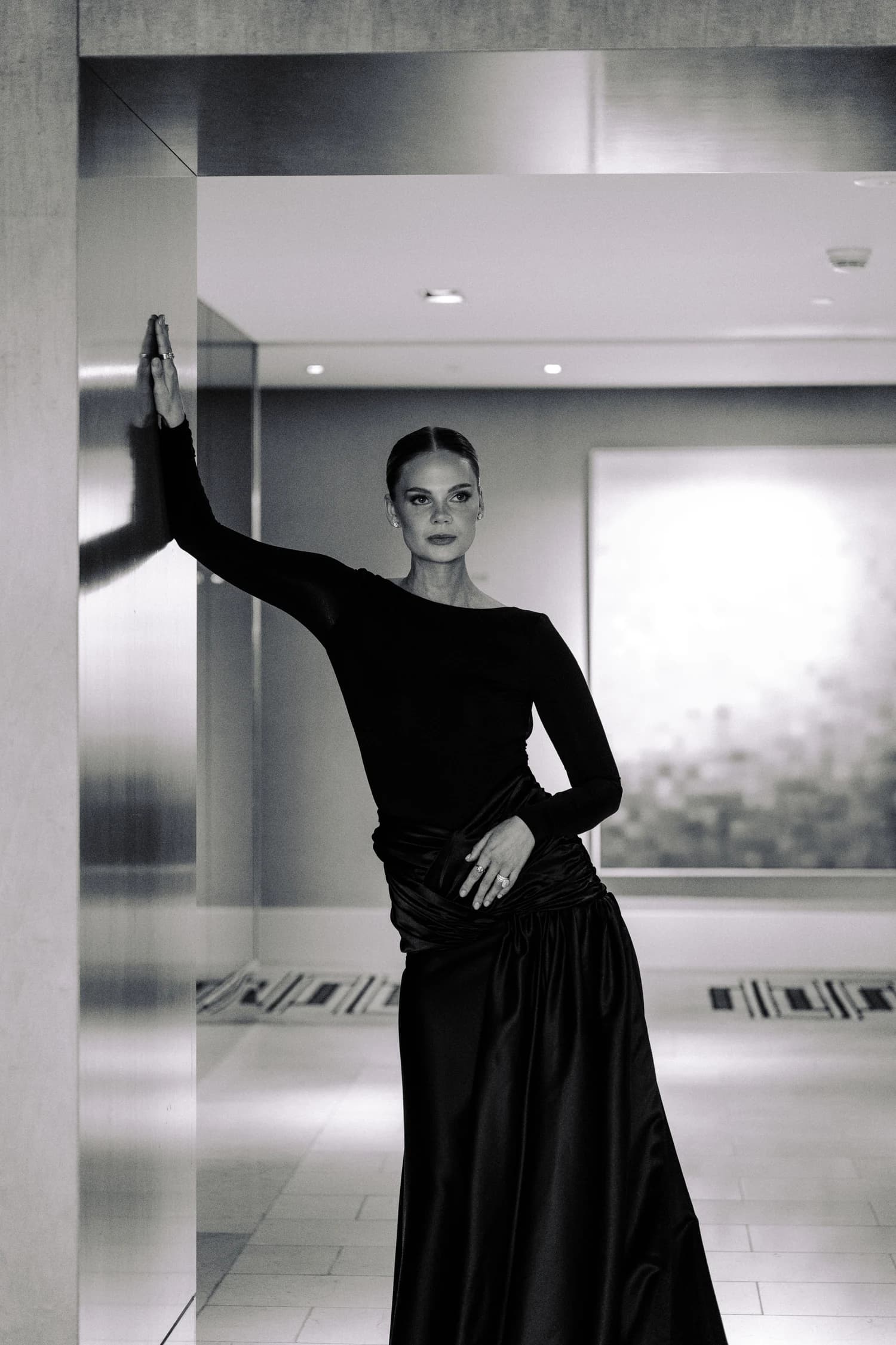 A woman posing in a floor-length black gown with one hand resting on a mirrored wall.