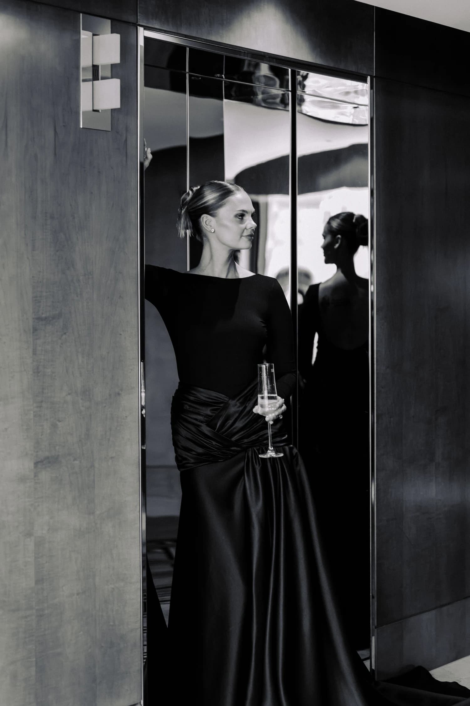 A woman in a floor-length black gown standing beside a mirrored wall.
