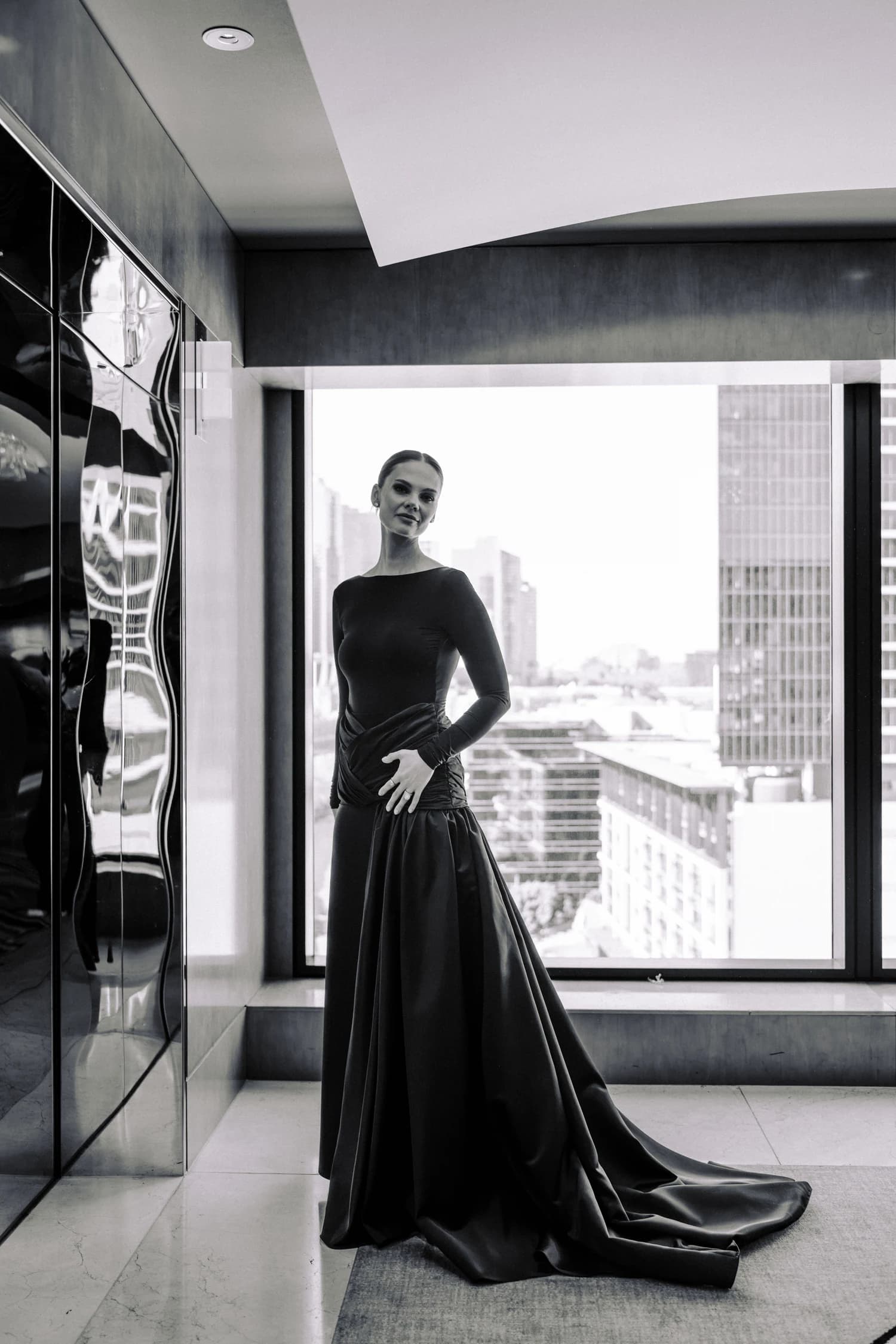 A woman standing beside a window in a black gown with its train spread across the carpet.