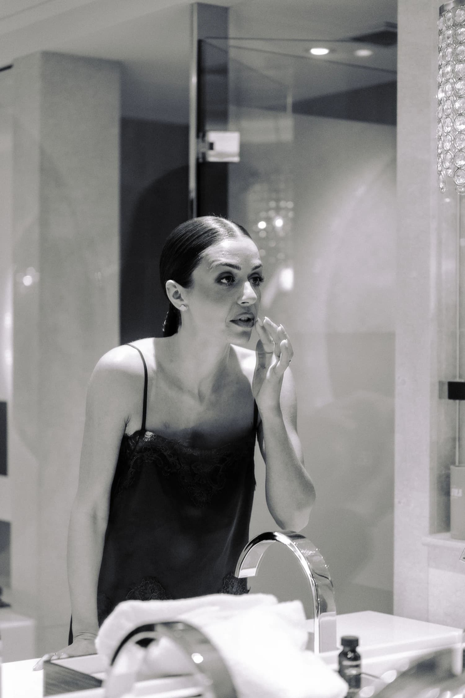A woman applying makeup at a bathroom mirror in a black lace camisole.