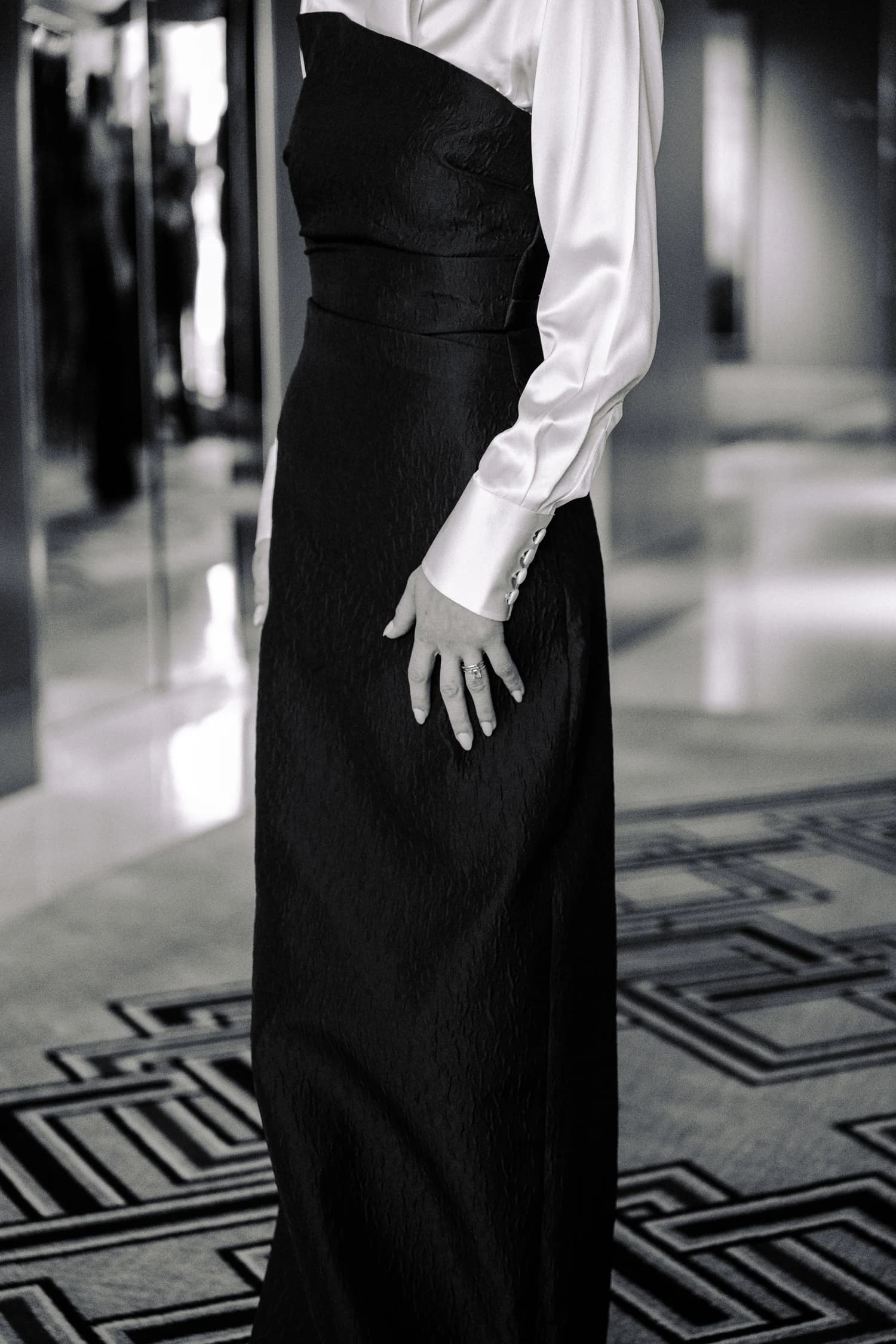 A satin shirt cuff and hand against a fitted black gown, with patterned carpet behind.