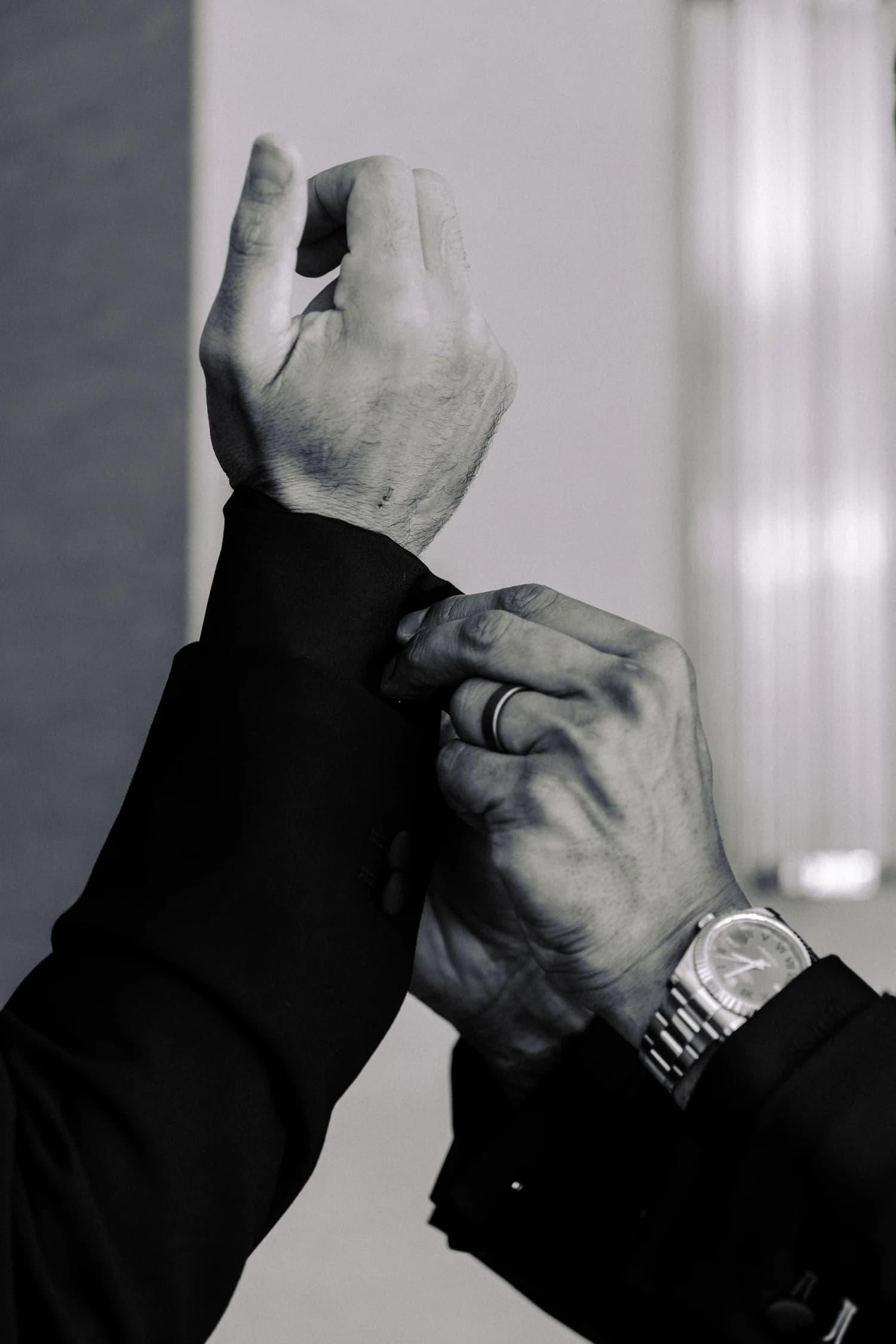 Hands fastening a black shirt cuff beside a metal wristwatch.