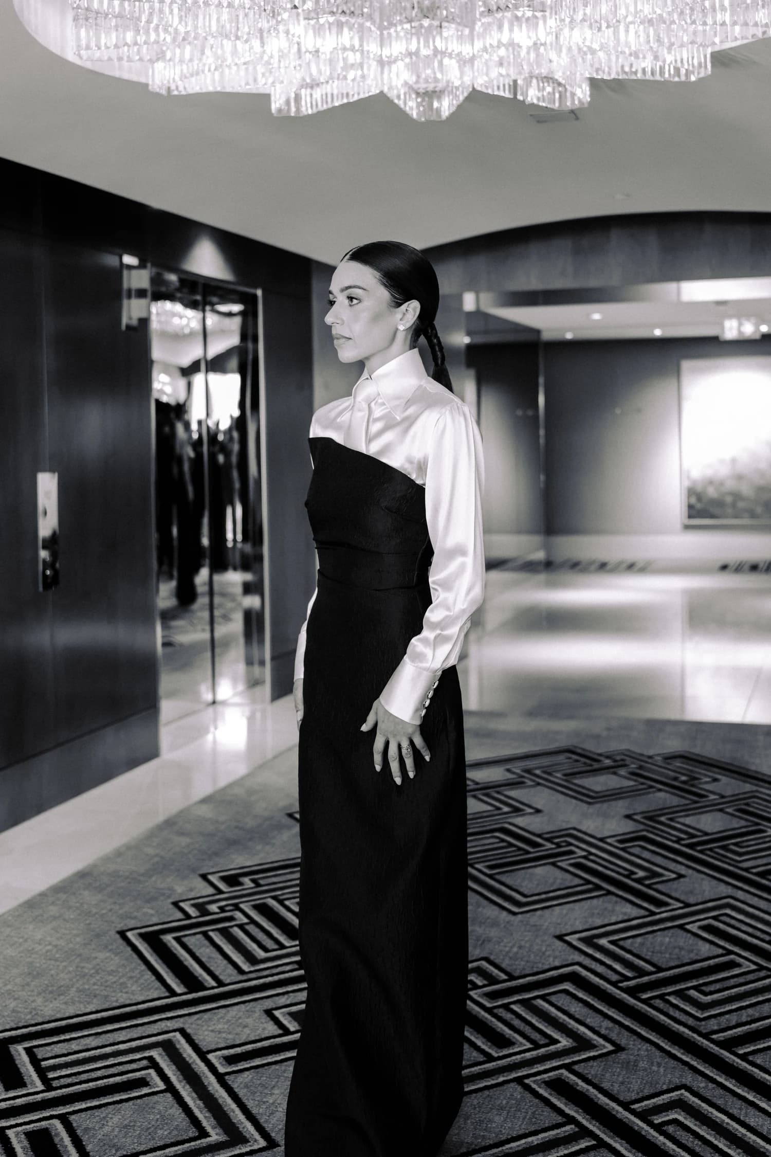 A full-length portrait of a woman in a white blouse and black gown beside a mirror.