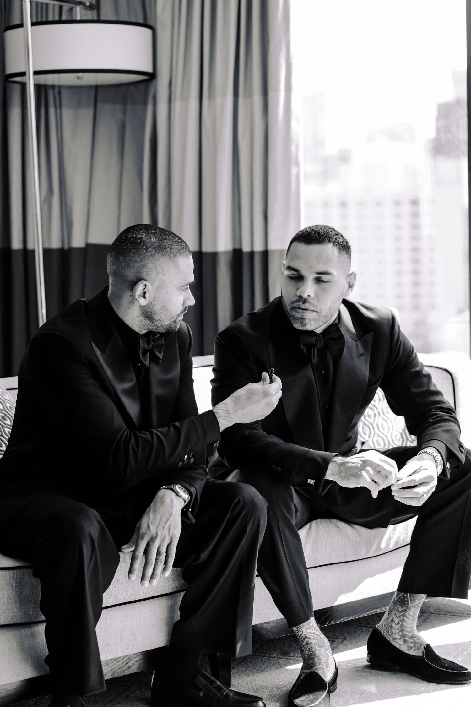 Two men in dark suits seated together on a sofa beside the dressing-room window.
