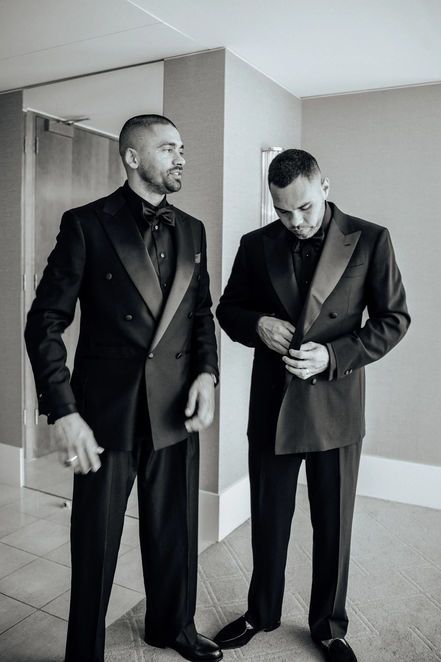 Two men standing in dark suits, one adjusting his shirt cuff.