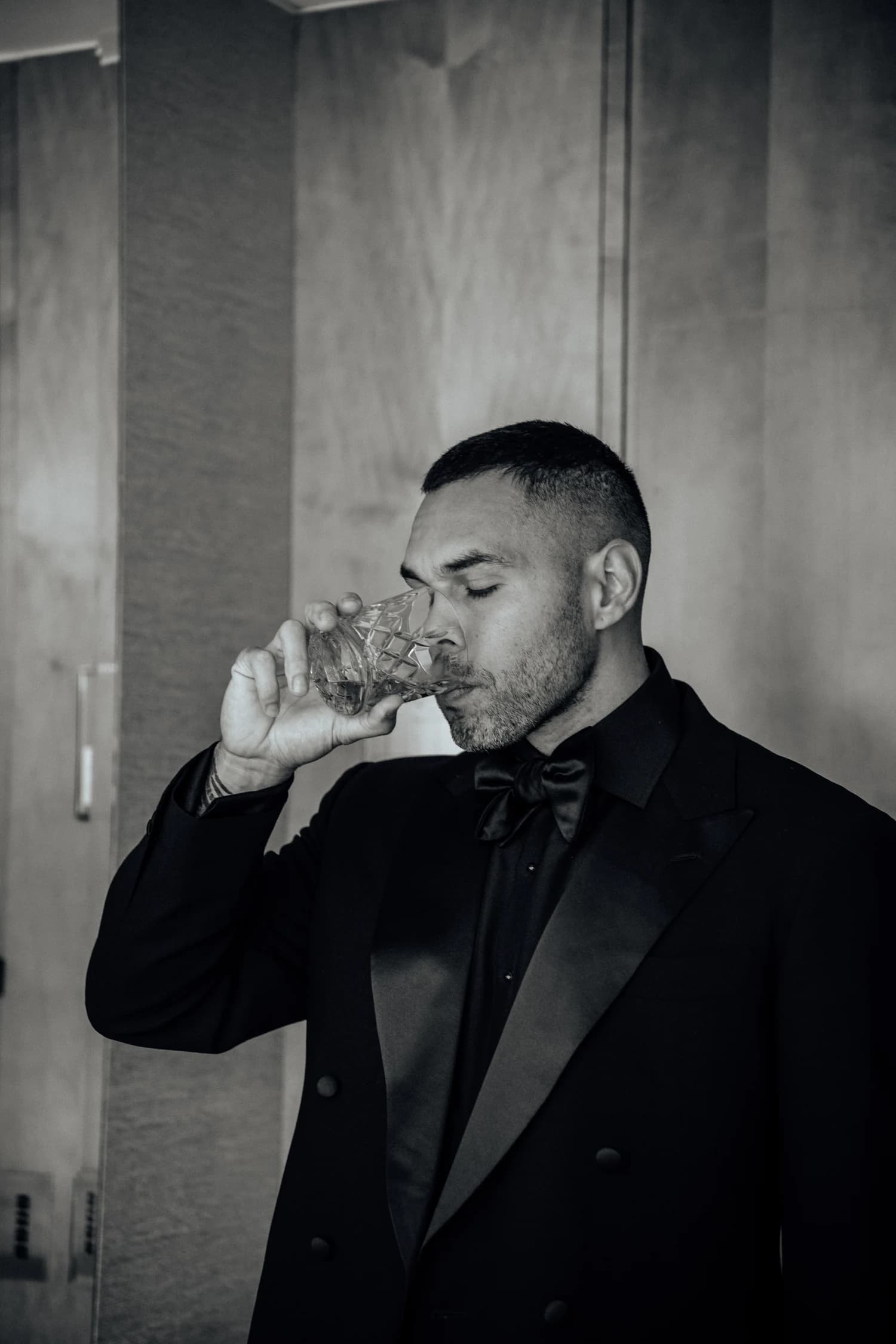 A man in a dark double-breasted suit and bow tie lifting a glass beside a timber doorway.