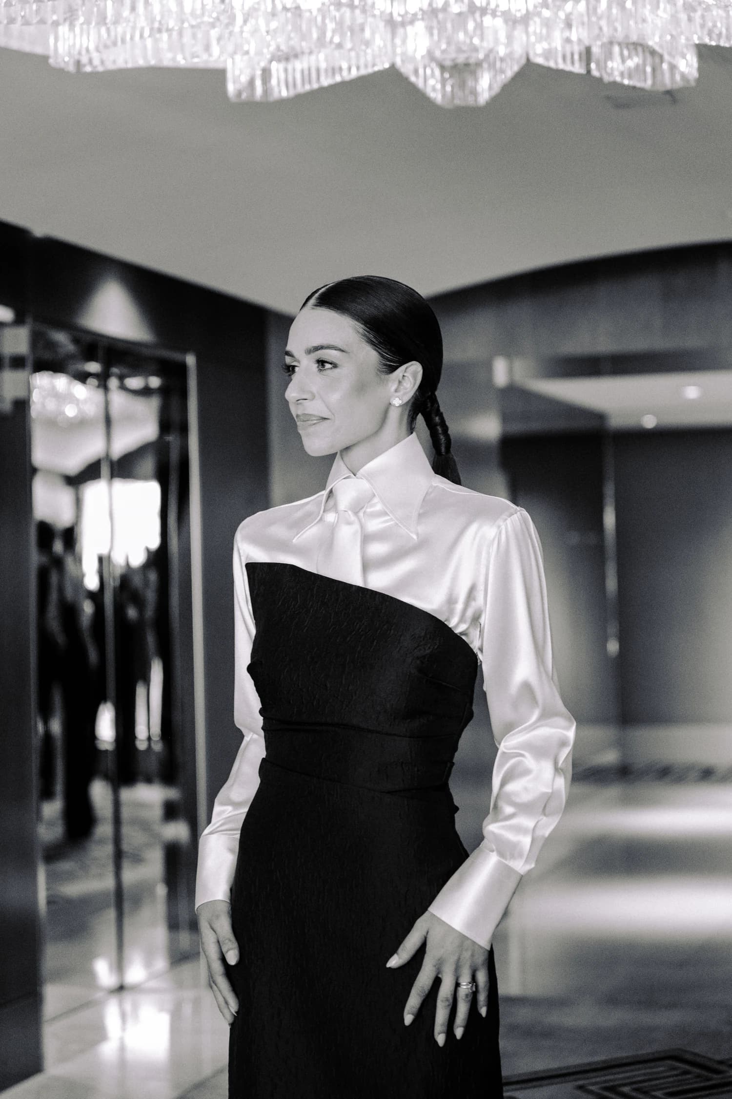 A woman wearing a white satin blouse with a fitted black gown, photographed beside a mirrored wall.