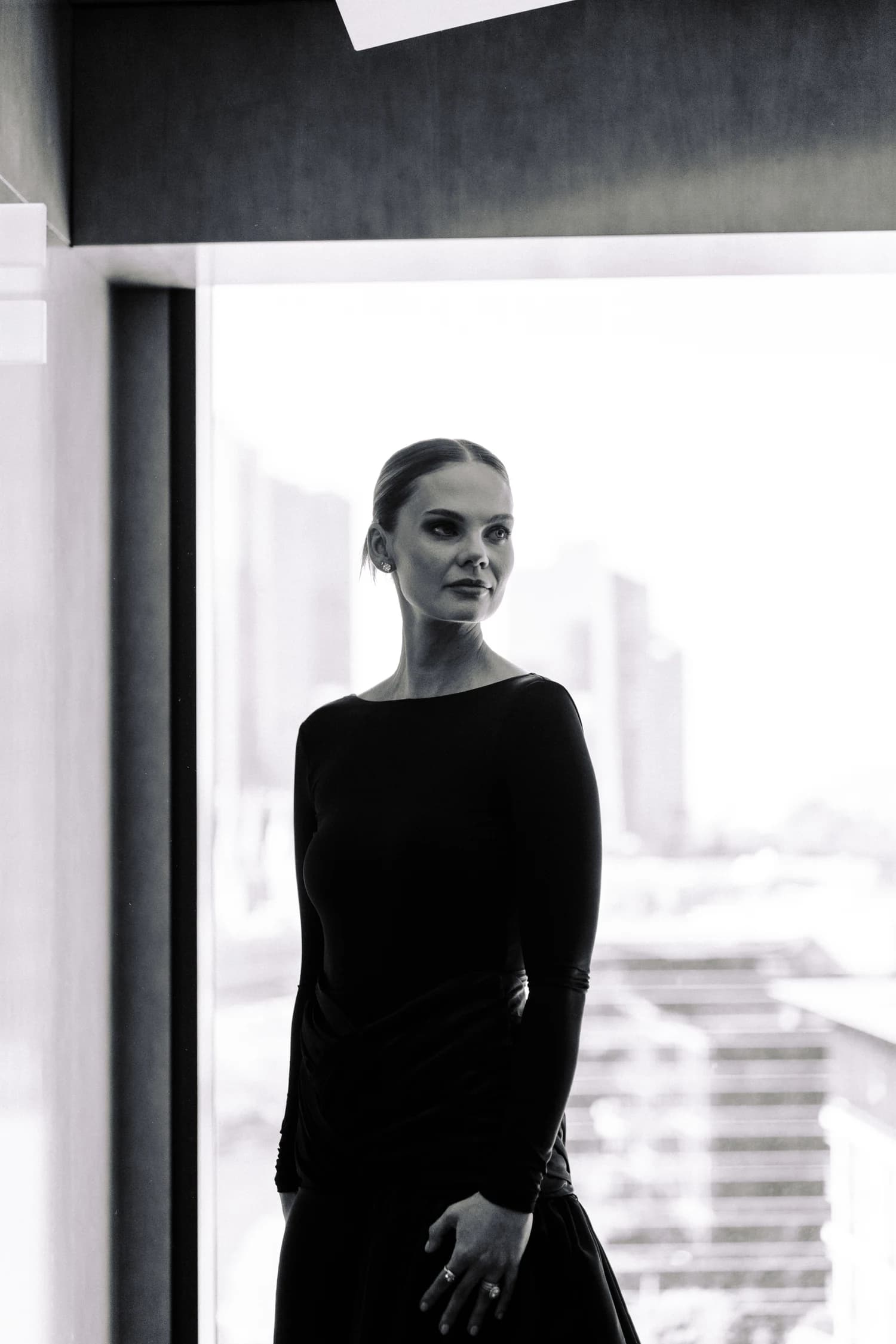 A woman in a black gown posing beside a bright dressing-room window.