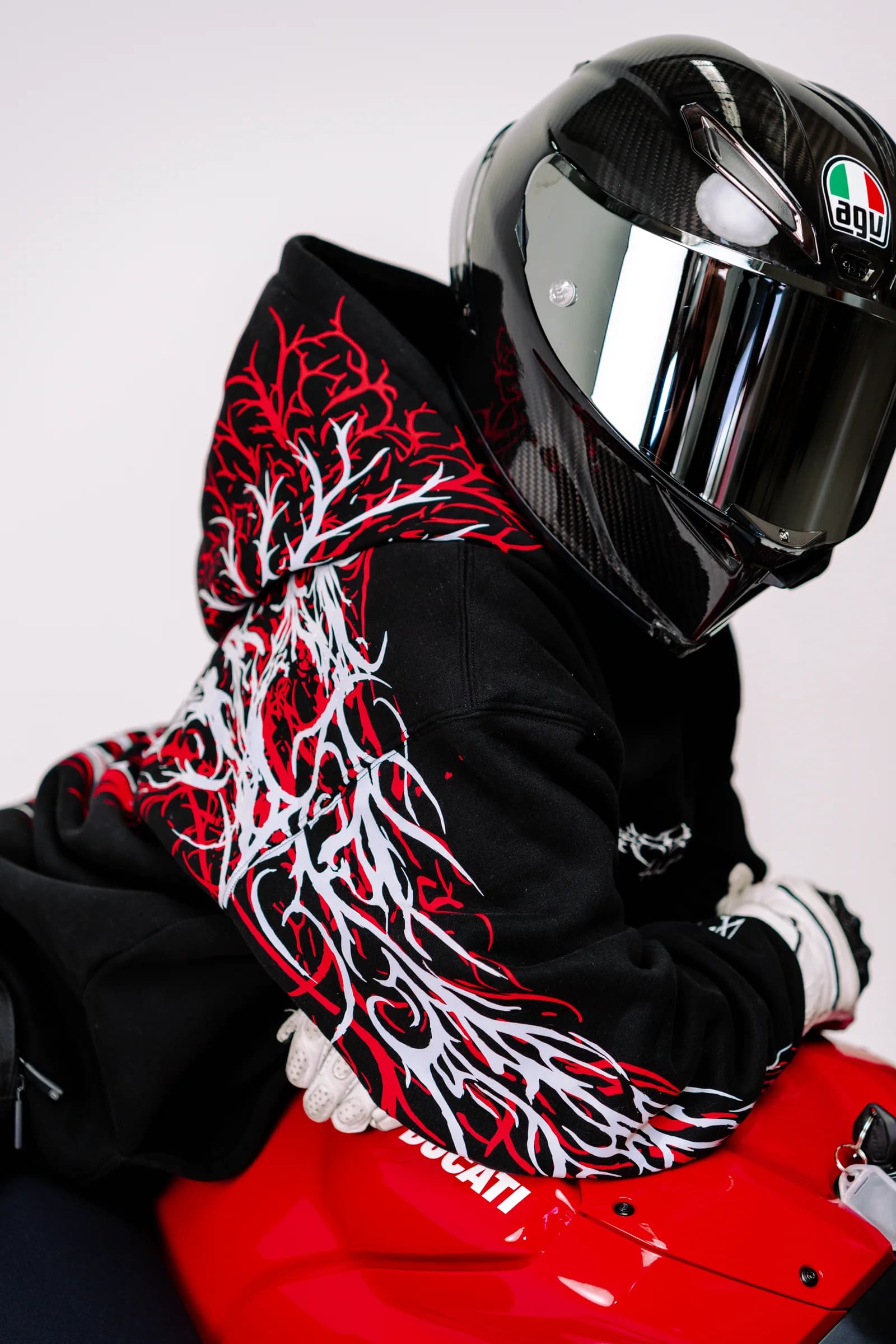 A helmeted model wearing a black 444racingco hoodie beside a red motorcycle in a white studio