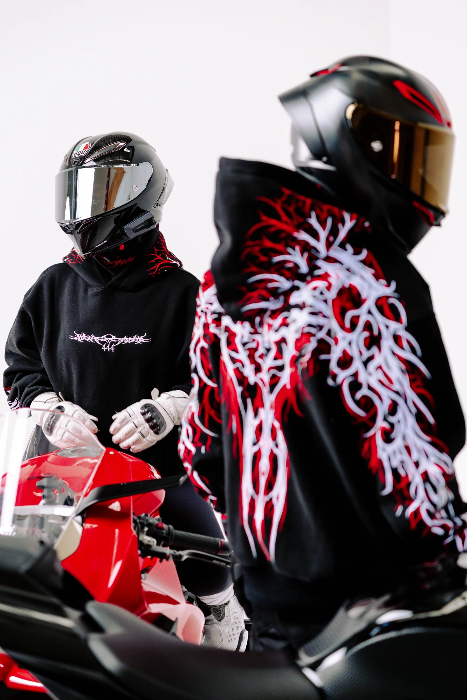 Two helmeted models in black hoodies beside a red motorcycle