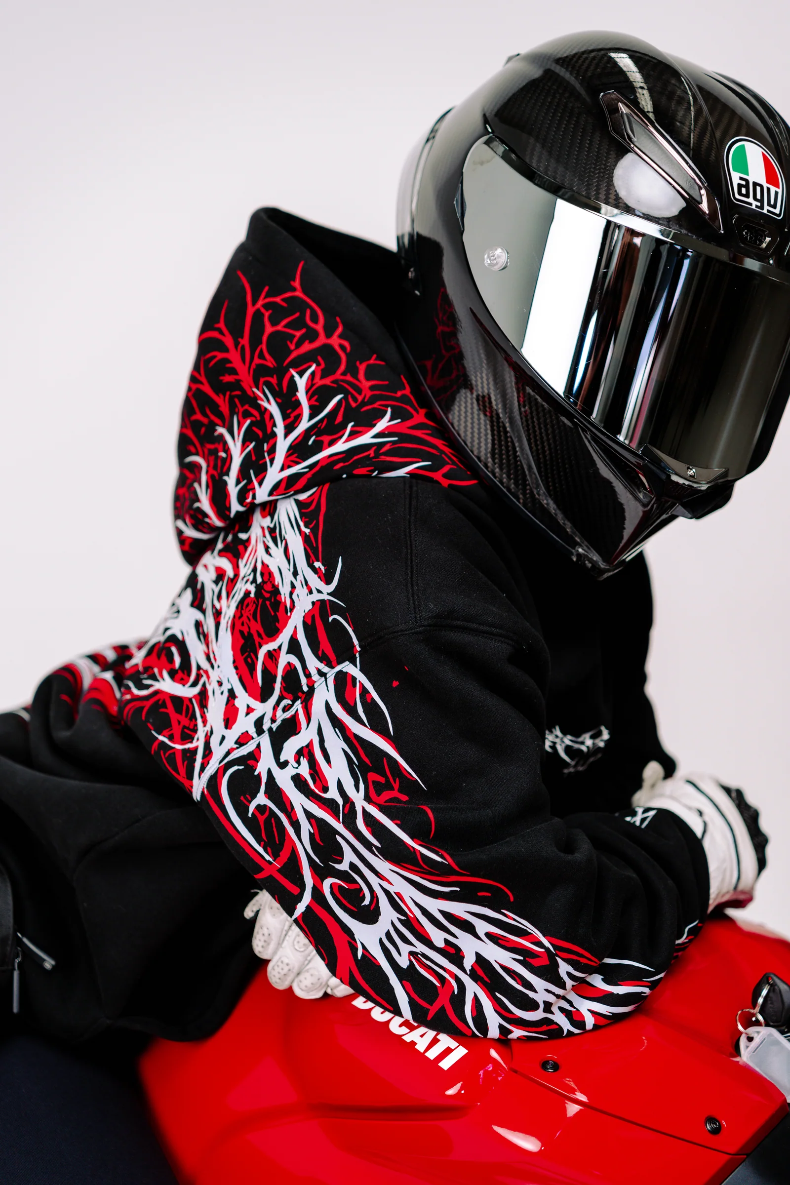 A helmeted model wearing a black 444racingco hoodie beside a red motorcycle in a white studio