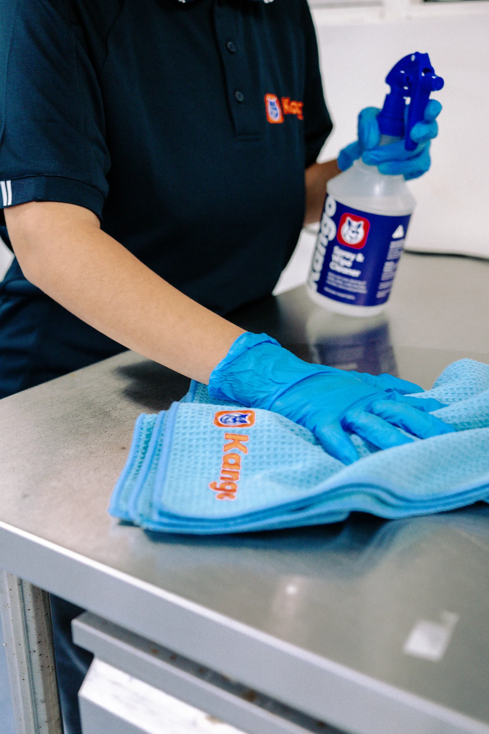 A gloved hand cleaning a stainless bench with Kango cloths and a spray bottle